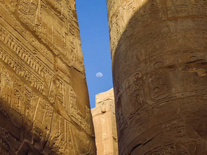 Carved columns of Karnak Temple framing a small Moon in a blue sky