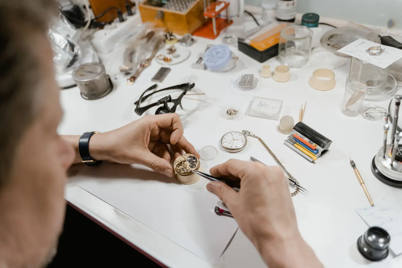 A watchmaker's hands at a white bench, lifting a part from a movement with tweezers, loupe and tools laid out around it